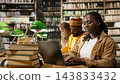 Student, laptop and books frame woman typing at wooden library table with classmates studying behind her. Coursework, digital literacy and academic preparation support learning through technology. 143833432