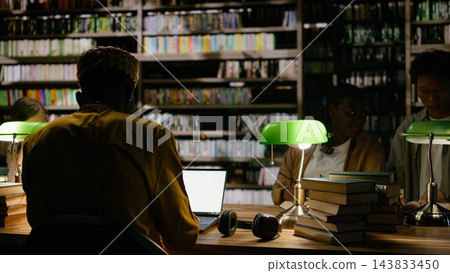 Adult learner, laptop and library frame rear view of man in yellow clothing working at computer near open books. Continuing education, research and digital skills shape study at communal table. Adult learner, laptop and library frame rear view of man in yellow clothing working at computer near open books. Continuing education, research and digital skills shape study at communal table. 143833450