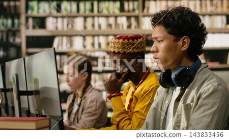 Student, headphones and computer as young man focuses on desktop monitor while classmates work nearby in library. Digital literacy, independent study and academic focus support computer learning. Student, headphones and computer as young man focuses on desktop monitor while classmates work nearby in library. Digital literacy, independent study and academic focus support computer learning. 143833456