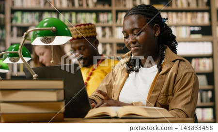 Student, laptop and book as smiling woman works on keyboard beside open reference text at library table. Digital learning, productivity and research connect academic reading with computer coursework. Student, laptop and book as smiling woman works on keyboard beside open reference text at library table. Digital learning, productivity and research connect academic reading with computer coursework. 143833458