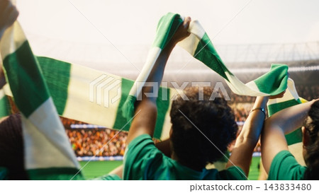 Soccer supporters, green scarves and raised arms fill stands as fans wave team colors above their heads. Fan culture, unity and crowd energy express collective enthusiasm during stadium match. Soccer supporters, green scarves and raised arms fill stands as fans wave team colors above their heads. Fan culture, unity and crowd energy express collective enthusiasm during stadium match. 143833480
