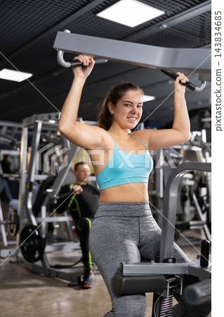 Young female training trains the muscles of the arms and press on simulator in gym. Sport concept Young female training trains the muscles of the arms and press on simulator in gym. Sport concept 143834685