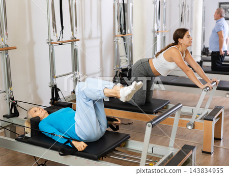 Focused elderly woman lying while doing Pilates tone exercise for back on reformer bed equipment in gym Focused elderly woman lying while doing Pilates tone exercise for back on reformer bed equipment in gym 143834955