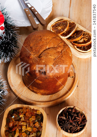 Whole Italian panettone on a bamboo tray, accompanied by candied fruits and dehydrated orange slices, set on a rustic wooden Christmas table 143837540