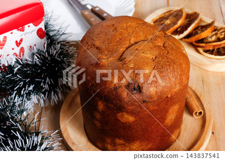 Whole Italian panettone on a bamboo tray, accompanied by candied fruits and dehydrated orange slices, set on an ornate, rustic wooden Christmas table 143837541