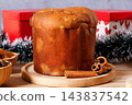 Whole Italian panettone on a round bamboo board, surrounded by candied fruits and dehydrated orange slices, set on a rustic wooden table 143837542