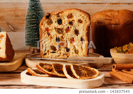 Traditional Christmas panettone, sliced and filled with candied fruit, surrounded by dehydrated orange slices on a rustic wooden table 143837545