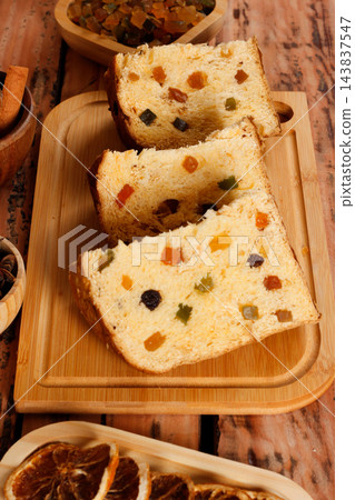 Slices of traditional Italian panettone filled with raisins and colorful candied fruits arranged on a bamboo board atop a rustic wooden Christmas table 143837547