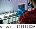 Worker wearing blue protective gloves inspecting glass bottles with beverage on conveyor belt inside beverage production factory plant for quality control process check 143838809