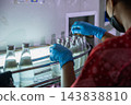 Worker wearing blue gloves and face mask operating production line in beverage factory, checking glass bottles filled with refreshing clean liquid during quality control process inside facility 143838810