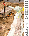 A wheelbarrow loaded with gray gravel stands next to a freshly dug trench lined with protective fabric at a busy landscaping site. 143838861