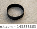 Under soft lighting, a black rubber drive belt rests flat on a gray concrete background. This mechanical component is crucial for machinery operation and shows a textured surface. 143838863