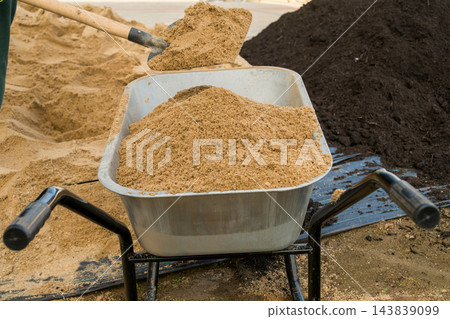 A shovel is actively used to fill a metal wheelbarrow with sand at a busy job site. Nearby, a large pile of sand reflects the ongoing landscaping work. 143839099