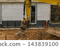 A yellow excavator is actively engaged in digging soil beside a building's foundation at a construction site. 143839100