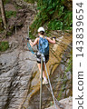 Back view of female climber crosses suspended cable bridge on Via Ferrata Murren-Gimmelwald, balancing above rocky gorge with helmet, harness and backpack during alpine adventure in Switzerland 143839654