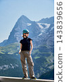 Young female traveler wearing hiking helmet and sunglasses stands at scenic viewpoint and looks across Swiss Alps, with massive Eiger mountain, glacier, green alpine slopes forming summer background 143839656