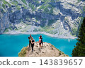 Group of hikers pauses on rocky overlook above vivid turquoise Oeschinensee, enjoying mountain scenery and taking photos during summer trekking trip near Kandersteg, Switzerland 143839657