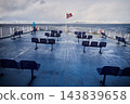 Wide view of winter ferry deck with lone woman near railing, Norwegian flag flying above empty passenger seats and snowy mountain coastline stretching across cold sea near Lofoten Islands 143839658