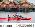 Happy kayaking group celebrates on calm water in Hamnoy, raising hands and paddles from two tandem kayaks with iconic red fishing cabins behind during wet winter weather in Lofoten, Norway 143839664