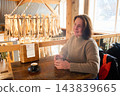 Smiling woman enjoys quiet coffee break in rustic restaurant in Reine, Lofoten, holding glass of water beside espresso while dried fish display creates distinctive Norwegian coastal atmosphere 143839665