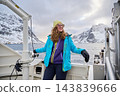 Young woman in sunglasses poses on boat deck during winter sightseeing trip near Reine, Lofoten Islands, Norway, with fjord and snow covered mountains creating dramatic Arctic backdrop 143839666