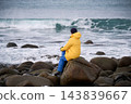 Woman sits on large boulder watching powerful waves of Norwegian Sea at Unstad Beach, Lofoten, Norway, surrounded by rocky winter shoreline during cold Arctic coastal weather 143839667