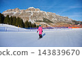 Female snowboarder rides across broad groomed Sellaronda ski slope with dramatic rocky Dolomites massif, conifer forest, safety fencing, sunny blue sky forming scenic winter resort landscape in Italy 143839670