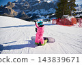 Female snowboarder takes smartphone selfie while kneeling on snowy slope in Dolomites, Italy, with snowboard beside her and dramatic alpine cliffs, resort terrain and bright winter scenery behind 143839671