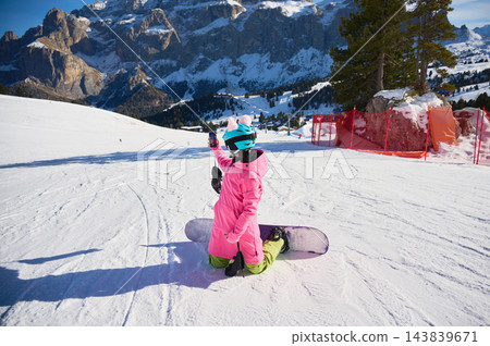 Female snowboarder takes smartphone selfie while kneeling on snowy slope in Dolomites, Italy, with snowboard beside her and dramatic alpine cliffs, resort terrain and bright winter scenery behind Female snowboarder takes smartphone selfie while kneeling on snowy slope in Dolomites, Italy, with snowboard beside her and dramatic alpine cliffs, resort terrain and bright winter scenery behind 143839671