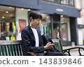 A male businessman in his twenties sitting on a bench and looking at his smartphone 143839846