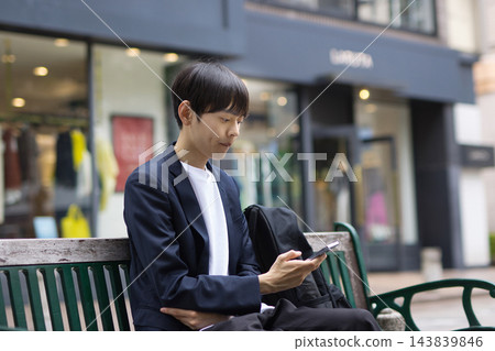 一位二十多歲的男性商人坐在長椅上看著他的智慧型手機 143839846