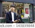 A male businessman in his twenties sitting on a bench and looking at his smartphone 143839848
