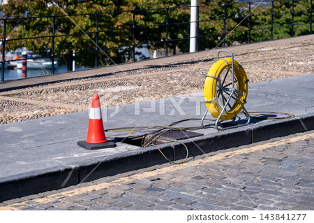 Yellow fiber optic cable reel on a metal stand placed on a concrete sidewalk next to an open utility manhole. Maintenance or telecommunication installation work place 143841277