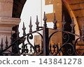 Decorative wrought iron gate spikes with intricate forged scrolls. Vintage rusted metal texture and ancient stone archway architecture in the blurred background 143841278