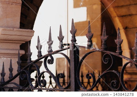 Decorative wrought iron gate spikes with intricate forged scrolls. Vintage rusted metal texture and ancient stone archway architecture in the blurred background 143841278