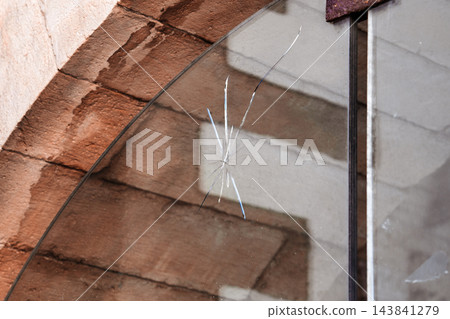 Crack and impact mark on a transparent glass window pane installed inside an old stone archway Crack and impact mark on a transparent glass window pane installed inside an old stone archway 143841279