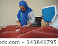 Woman tailor measures and marks red fabric with chalk and ruler beside a sewing machine in a bright home tailoring workshop 143841795