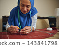 Sewing woman works red fabric by hand with a needle and thread beside measuring tape and tailoring tools in a textile workshop 143841798