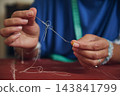 Detailed hands untangle white thread and hold a sewing needle above rust-colored fabric during handmade garment construction indoors 143841799