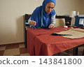 Woman tailor sews rust-colored fabric by hand with needle and thread at a worktable during handmade garment making indoors 143841800