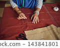 Tailor sews and pins rust-red fabric by hand while preparing a garment pattern at a bright creative home fashion workshop table 143841801