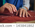 Focused hands sew rust-colored fabric with a needle and white thread during detailed hand stitching in a bright tailoring workshop 143841802
