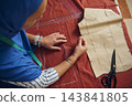 Woman tailor sews rust-red fabric by hand with needle, thread, patterns and measuring tape in a bright indoor home tailoring workshop 143841805