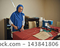 Woman tailors fabric with scissors beside a sewing machine in a home tailoring workshop, creating handmade clothing from garment patterns 143841806