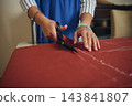Woman tailor cuts red fabric with scissors along chalk lines while preparing handmade clothing in a focused home tailoring workshop 143841807