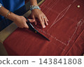 Tailor carefully cuts rust-red fabric with scissors along chalk pattern lines while making handmade clothing in a home sewing workshop 143841808