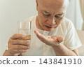 Senior man taking medication with a glass of water at home for health management. 143841910