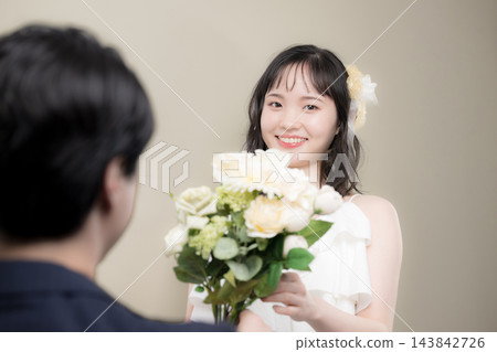 婚禮上,一位年輕的日本新娘穿著婚紗,手捧白色花束,幸福地對著新郎微笑。 婚禮上,一位年輕的日本新娘穿著婚紗,手捧白色花束,幸福地對著新郎微笑。 143842726