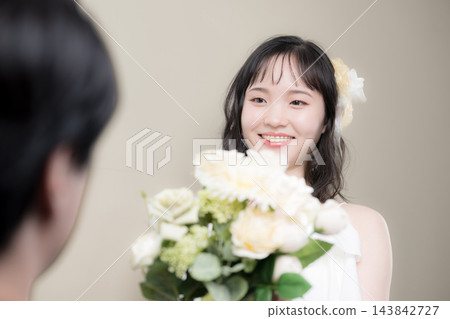 婚禮上,一位年輕的日本新娘穿著婚紗,手捧白色花束,幸福地對著新郎微笑。 婚禮上,一位年輕的日本新娘穿著婚紗,手捧白色花束,幸福地對著新郎微笑。 143842727