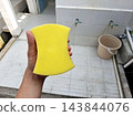 Hand holding ergonomic yellow sponge against outdoor tiled floor background. Tool used for effective scrubbing cleaning washing dishes and surfaces. 143844076